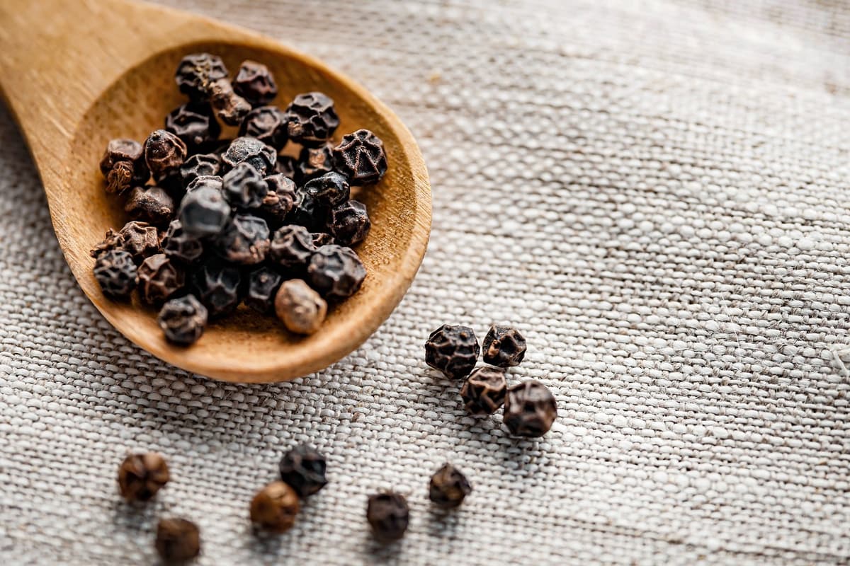 A wooden spoon heaped with dried black peppercorns, more scattered on a woven cloth